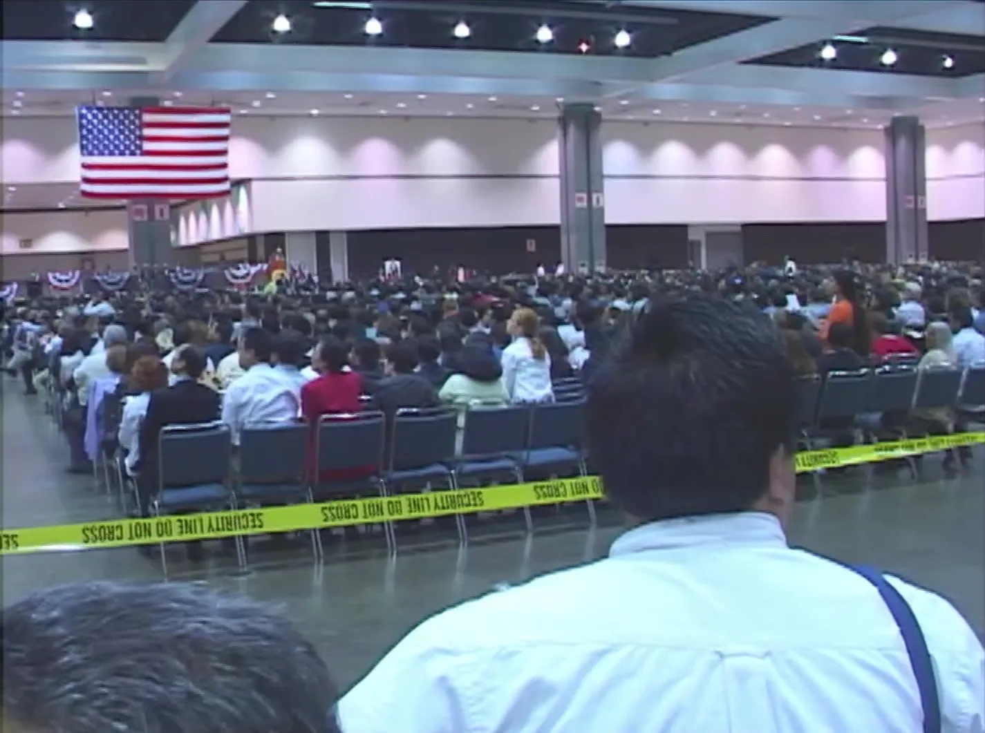 Hundreds of people seated in a convention center, seen from behind, with an American flag hanging at the front of the room.