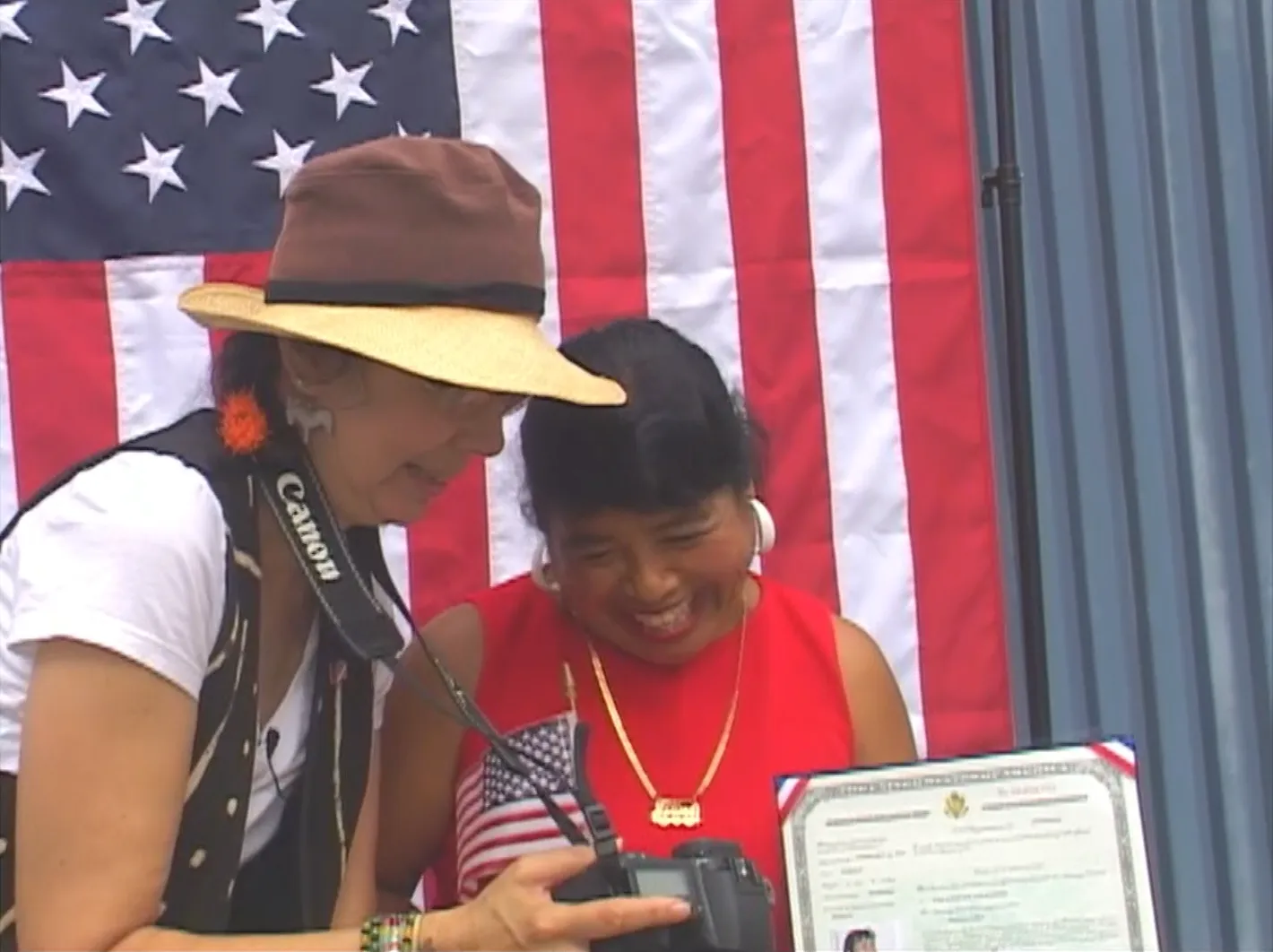 A woman on the left holds up a camera monitor to show a photo to a woman on the right. Both are smiling. The woman on the left holds a small American flag and a naturalization certificate, and there’s a large American flag in the background.