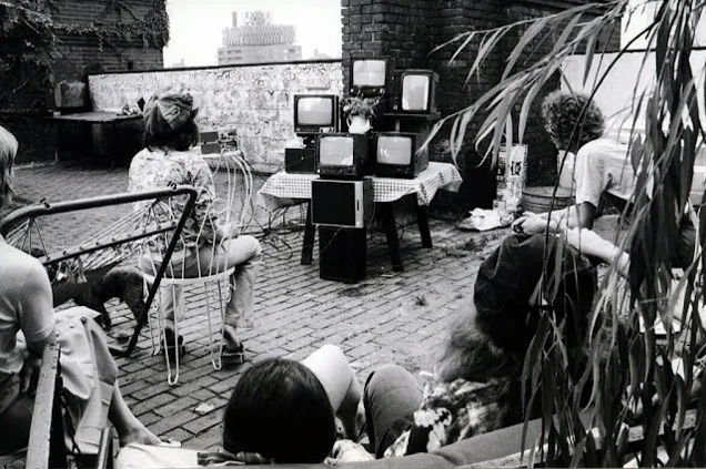 Black and white photograph of a group of people sitting on a rooftop looking at a series of video monitors stacked on a table.
